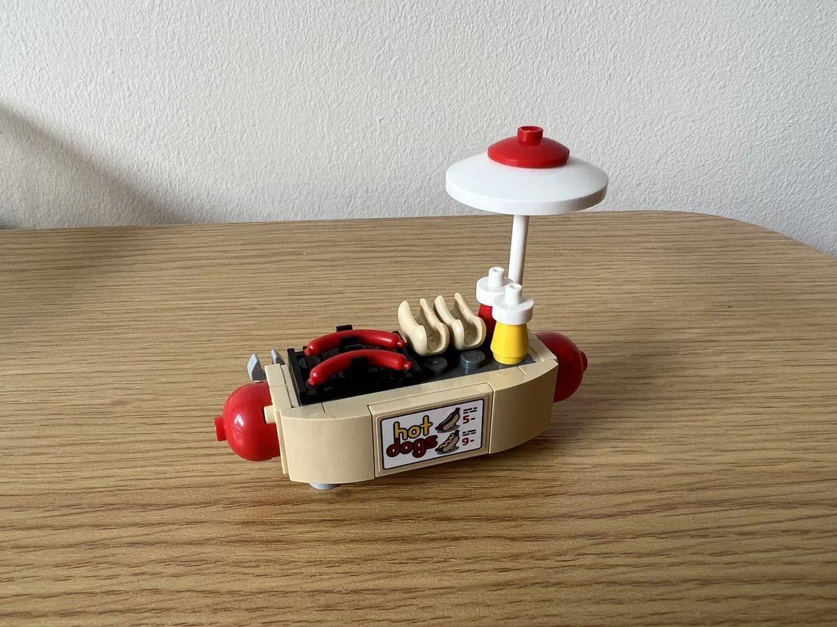 A lego hot dog cart shaped like a giant hot dog with the sausage poking out at each end. On top are two sausages and two buns, and a ketchup and mustard bottle.