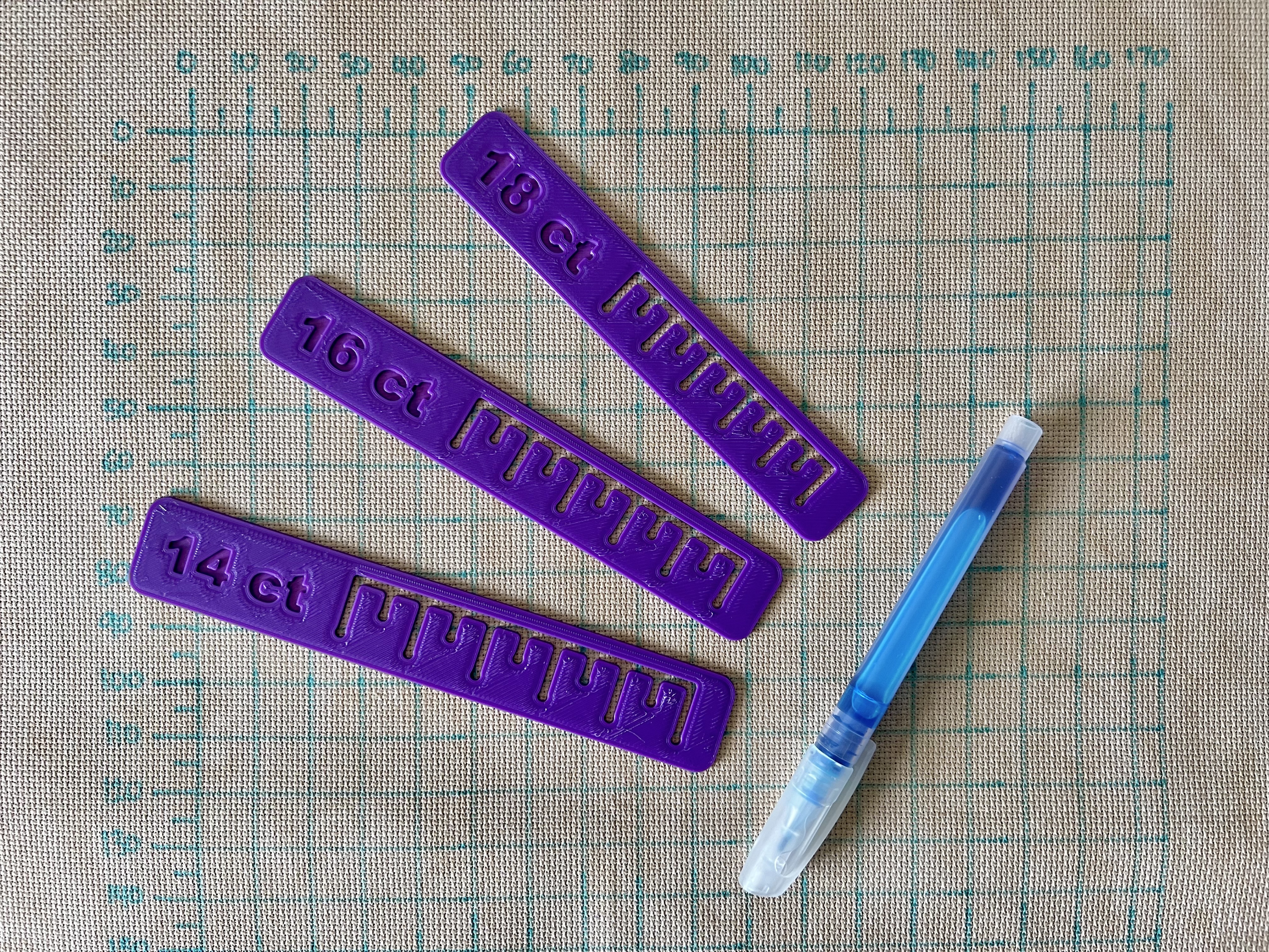 A piece of brown cross stitch fabric filling the frame with a grid drawn on in blue. On top of the fabric are three purple rulers and a blue pen.