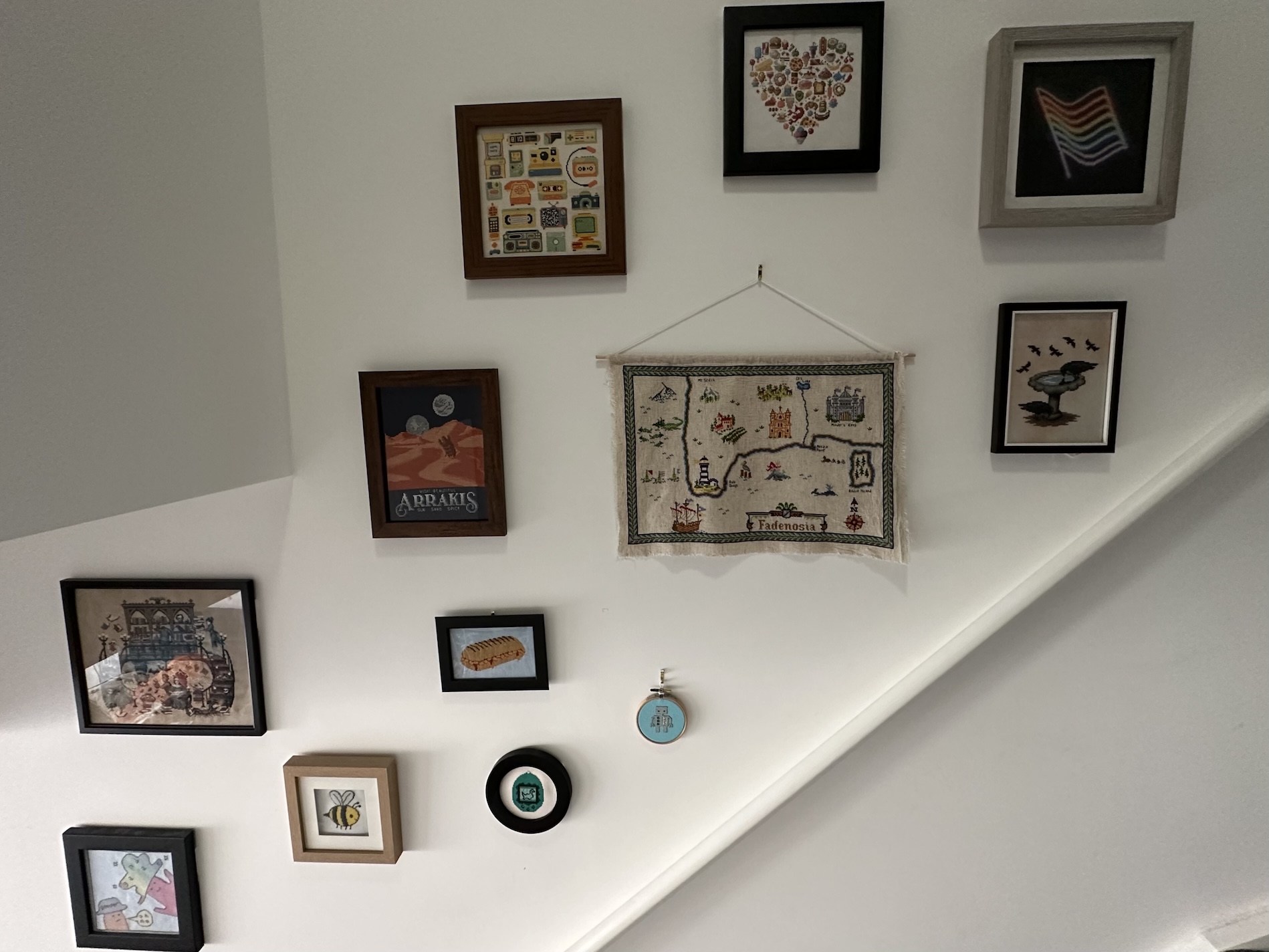 A set of twelve framed cross stitches hung on the wall above some stairs. All the images have different themes, and they're hung randomly on the wall rather than being aligned with one another.